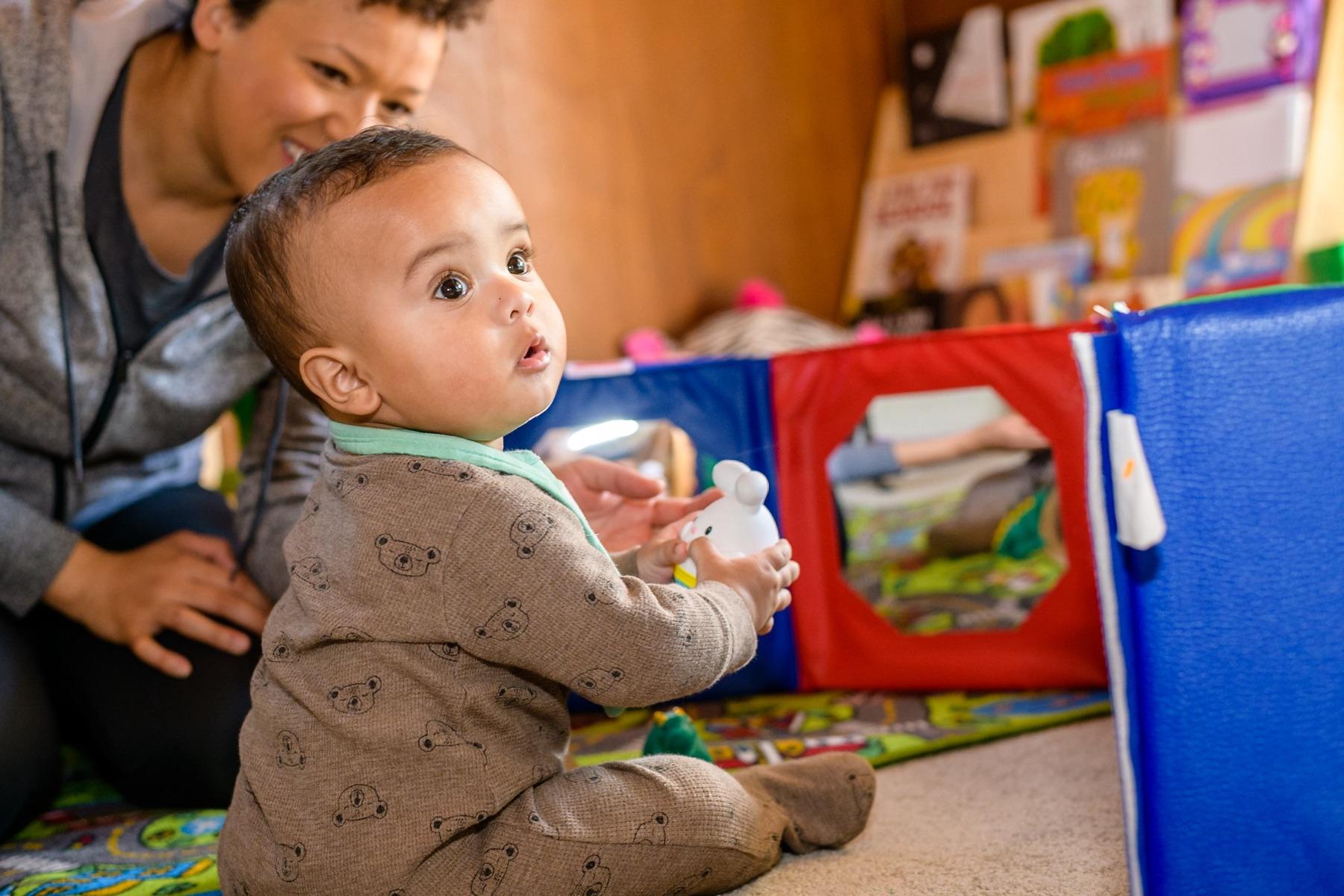 A baby sits on a play mat holding a toy while a caregiver smiles nearby